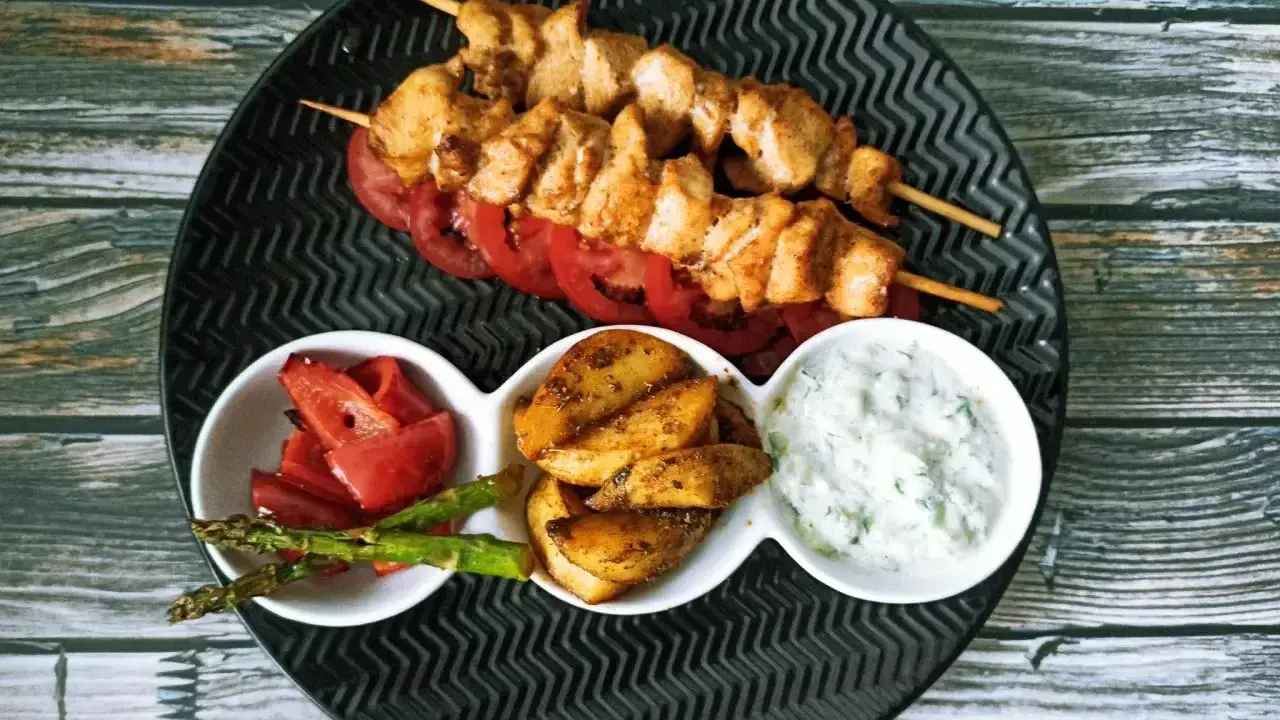 Dwa szaszłyki souvlaki z kurczaka na talerzu z pomidorami, ziemniakami, szparagami i sosem.