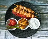 Dwa szaszłyki souvlaki z kurczaka na talerzu z pomidorami, ziemniakami, szparagami i sosem.