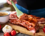 Pędzel rozprowadza aromatyczną marynatę do żeberek na surowym mięsie. Obok czosnek, pomidorki i papryczki chili.