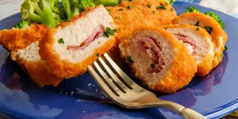Kotlet po szwajcarsku, panierowany, z szynką w środku, podany z brokułami na niebieskim talerzu. Złoty widelec czeka na pierwszy kęs.