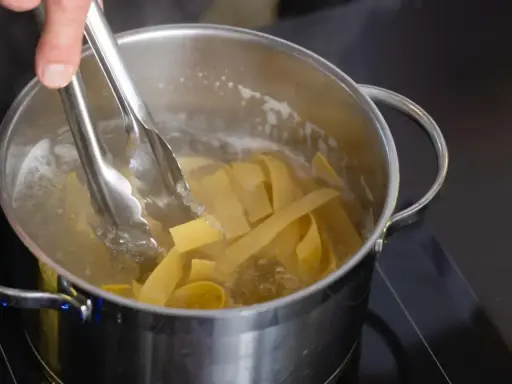 Szczypce do makaronu wkładają szerokie wstążki makaronu pełnoziarnistego do gotującej się wody w garnku.