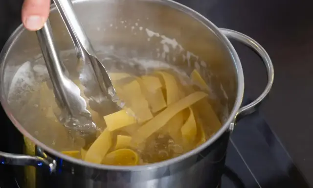 Szczypce do makaronu wkładają szerokie wstążki makaronu pełnoziarnistego do gotującej się wody w garnku.
