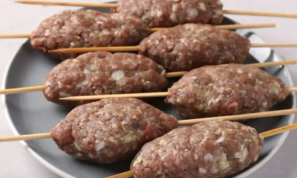 Siedem surowych kofta kebab na patyczkach, ułożonych na szarym talerzu, gotowych do grillowania.