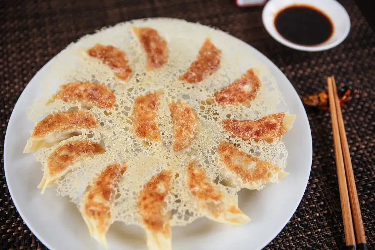 Złociste pierożki gyoza ułożone na talerzu, z chrupiącą, koronkową panierką. Obok miseczka sosu sojowego i pałeczki.