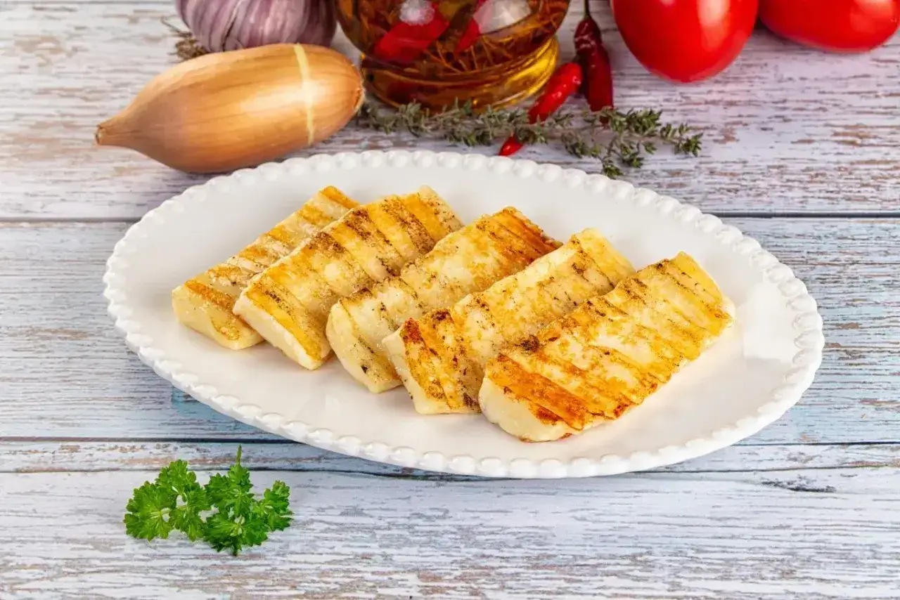Pięć kawałków grillowanego sera halloumi na białym talerzu, z dodatkami w tle: czosnek, cebula, pomidory, chili i zioła.