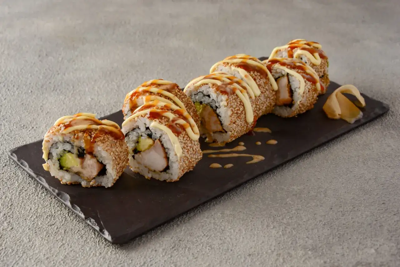 Siedem kawałków sushi z kurczakiem, obtoczonych w sezamie, polanych majonezem i sosem teriyaki, na ciemnej desce.